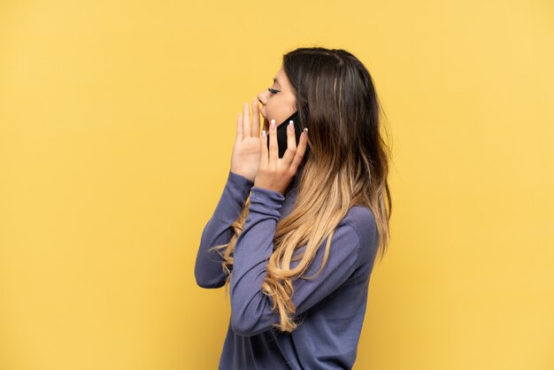 Young Russian girl using mobile phone isolated on yellow background shouting with mouth wide open to the side