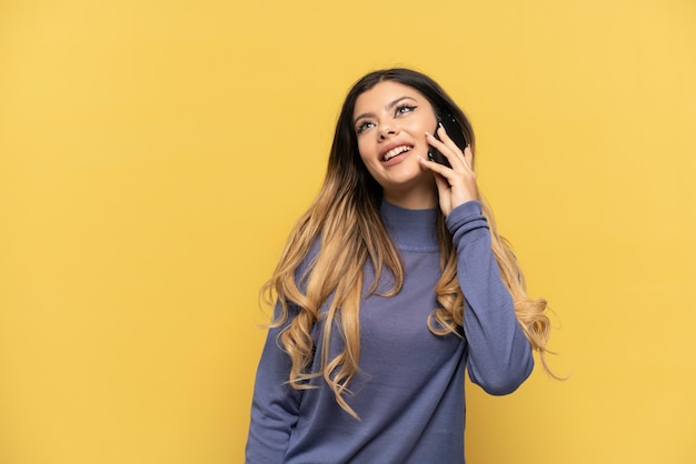 Young Russian girl using mobile phone isolated on yellow background thinking an idea while looking up