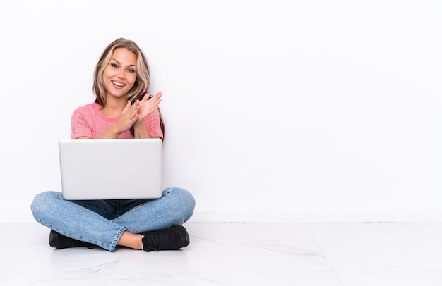 Young Russian girl with a laptop sitting on the floor isolated on white background applauding after presentation in a conference