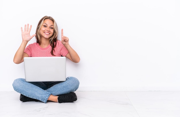 Young Russian girl with a laptop sitting on the floor isolated on white background counting six with fingers