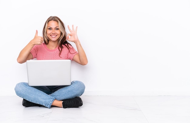 Young Russian girl with a laptop sitting on the floor isolated on white background showing ok sign and thumb up gesture