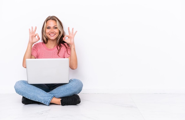 Photo young russian girl with a laptop sitting on the floor isolated on white background showing an ok sign with fingers