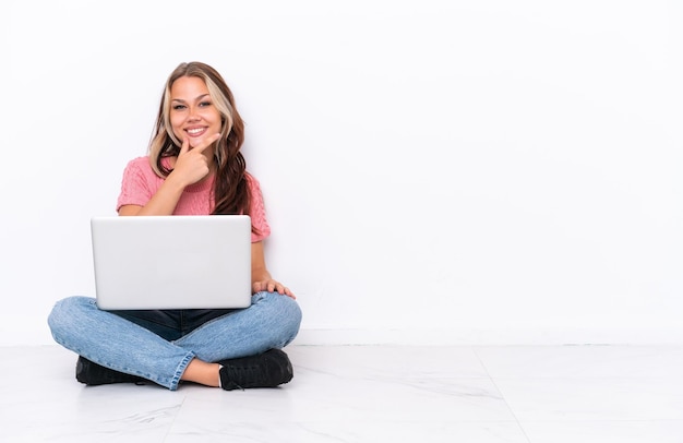 Giovane ragazza russa con un computer portatile seduto sul pavimento isolato su sfondo bianco sorridente