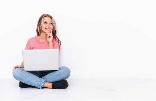 Young Russian girl with a laptop sitting on the floor isolated on white background thinking an idea while looking up