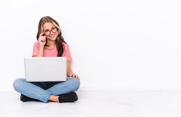 Young Russian girl with a laptop sitting on the floor isolated on white background with glasses and happy