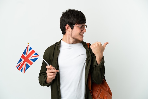 Молодой русский мужчина держит флаг Соединенного Королевства на белом фоне, указывая в сторону, чтобы представить продукт
