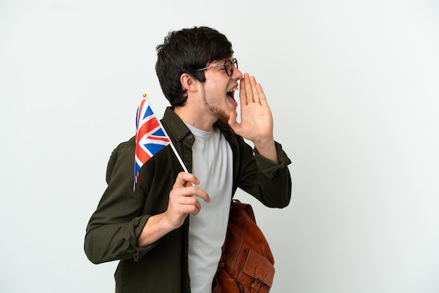 Young Russian man holding an United Kingdom flag isolated on white background shouting with mouth wide open to the side