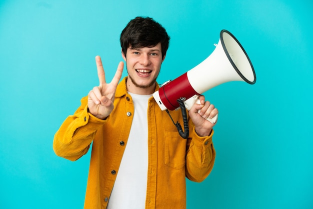 Young Russian man isolated on blue background holding a megaphone and smiling and showing victory sign