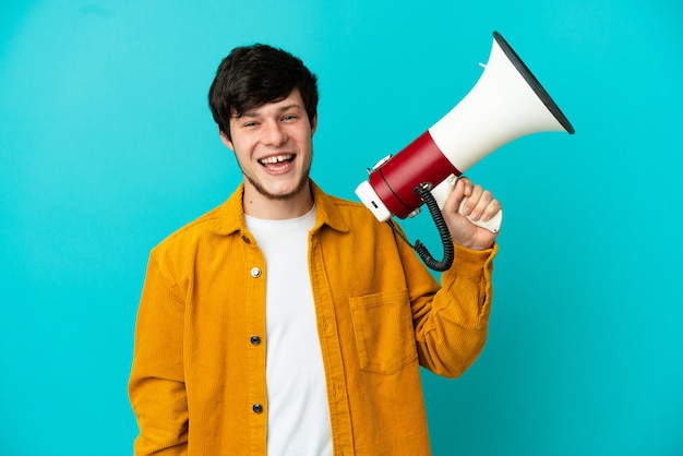 Young Russian man isolated on blue background holding a megaphone and smiling