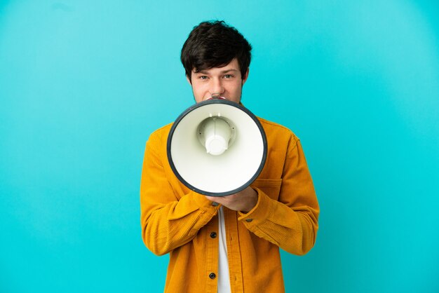 Young Russian man isolated on blue background shouting through a megaphone to announce something