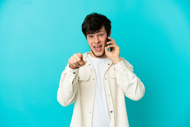 Young Russian man using mobile phone isolated on blue background surprised and pointing front