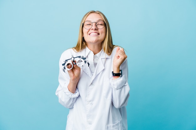 Giovane donna all'ottico optometrista russo sul pugno di sollevamento blu, sentendosi felice e di successo. concetto di vittoria.