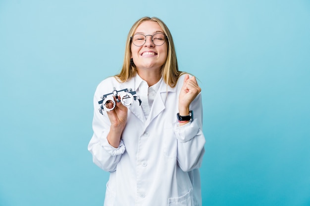 Young russian optometrist woman on blue raising fist, feeling happy and successful. Victory concept.