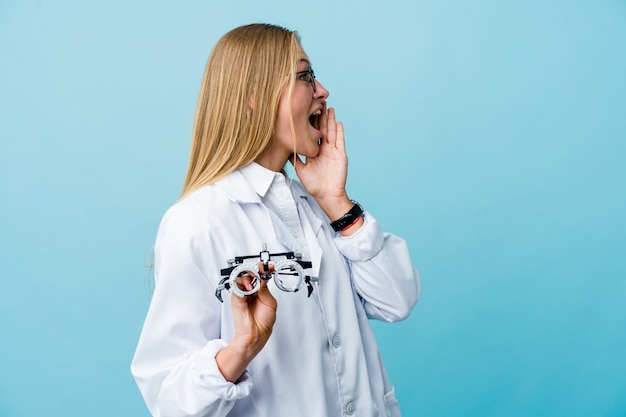 Young russian optometrist woman on blue shouting and holding palm near opened mouth.