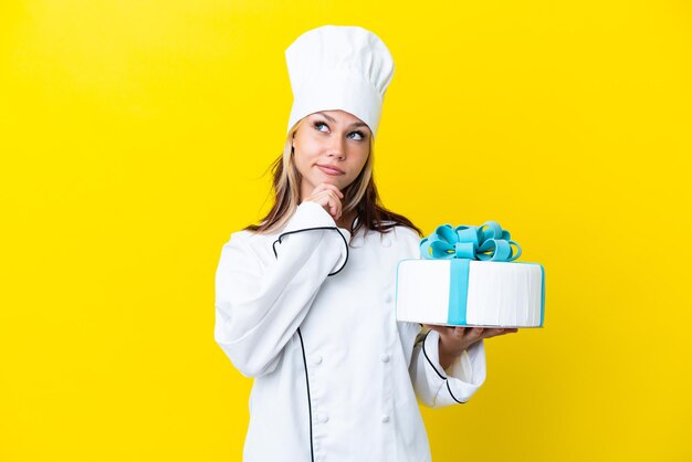 Giovane donna russa del pasticcere con una grande torta isolata su sfondo giallo e alzando lo sguardo