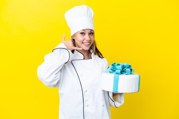 Young Russian pastry chef woman with a big cake isolated on yellow background making phone gesture. Call me back sign