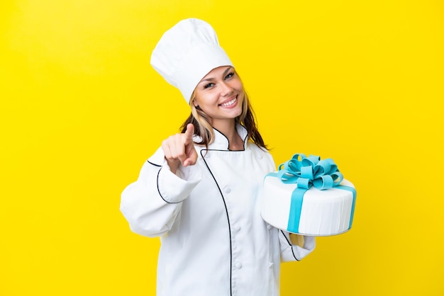 Young russian pastry chef woman with a big cake isolated on yellow background pointing front with happy expression