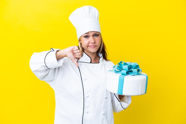 Young Russian pastry chef woman with a big cake isolated on yellow background showing thumb down with negative expression