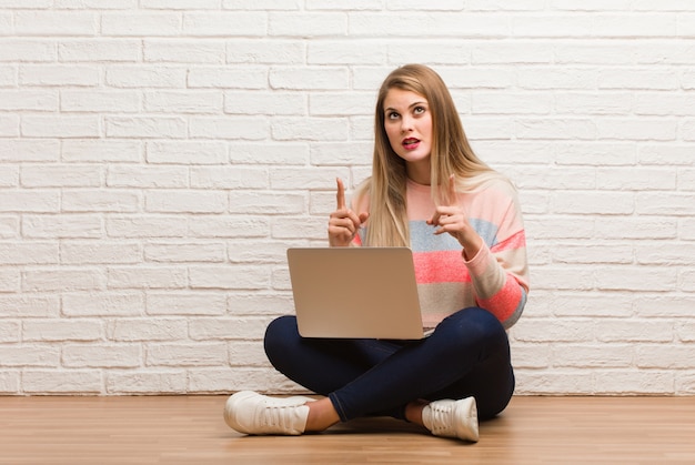 Young russian student woman sitting surprised pointing up to show something