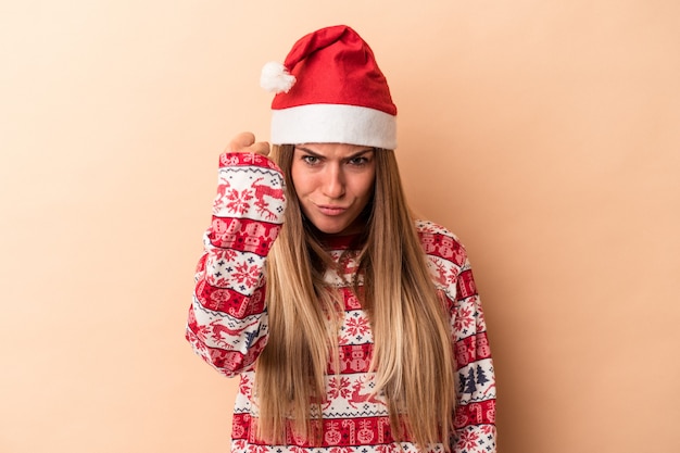 カメラに拳、攻撃的な表情を示すベージュの背景に分離されたクリスマスを祝う若いロシアの女性。