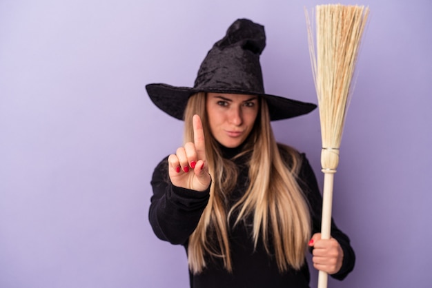 Фото Молодая русская женщина, замаскированная под ведьму, держит метлу, изолированную на фиолетовом фоне, показывая номер один пальцем.