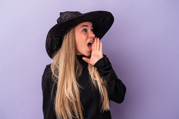 Young russian woman disguised as a witch celebrating halloween isolated on purple background shouting and holding palm near opened mouth.