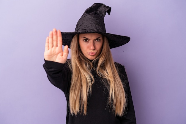 Молодая русская женщина, замаскированная под ведьму, празднует Хэллоуин, изолированную на фиолетовом фоне, стоя с протянутой рукой, показывая знак остановки, мешающий вам.