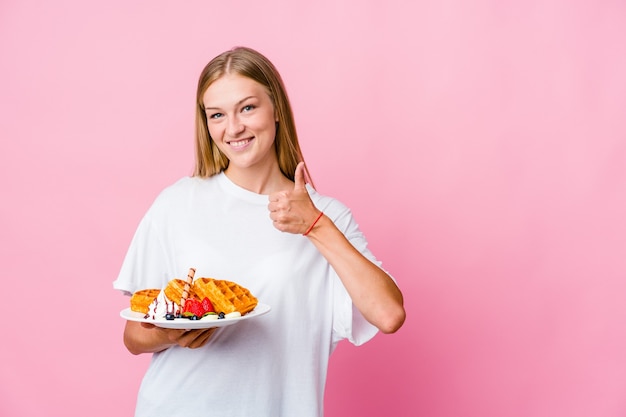 Молодая русская женщина ест изолированные вафли, улыбаясь и поднимая палец вверх