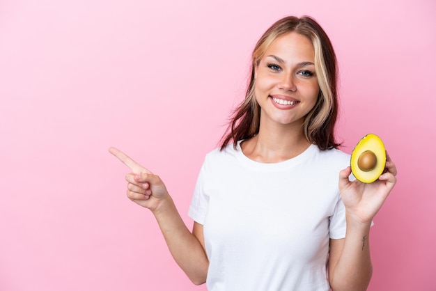 Young Russian woman holding avocado isolated on pink background pointing finger to the side