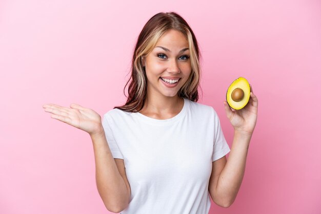 Young Russian woman holding avocado isolated on pink background with shocked facial expression