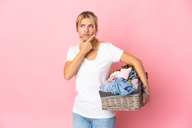 Young Russian woman holding a clothes basket isolated on pink background having doubts