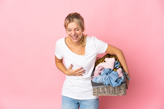 Young Russian woman holding a clothes basket isolated on pink background smiling a lot