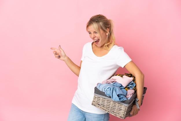 Young Russian woman holding a clothes basket isolated on pink wall pointing finger to the side and presenting a product