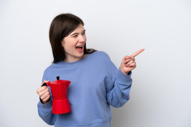 Молодая русская женщина держит кофейник на белом фоне, указывая пальцем в сторону и представляя продукт