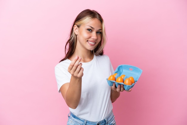 Young Russian woman holding eggs isolated on pink background making money gesture