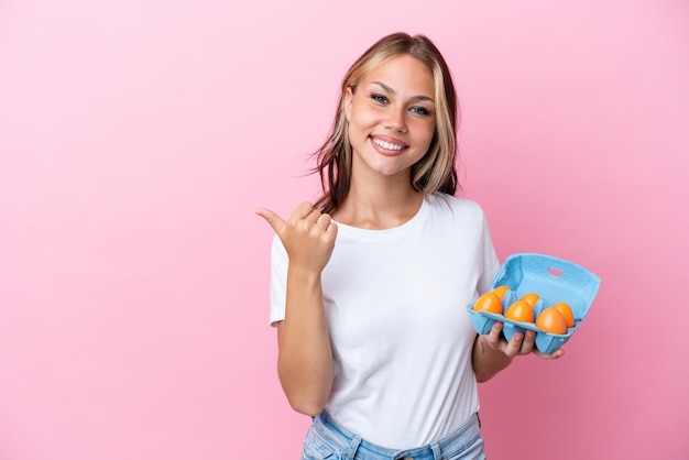 Young Russian woman holding eggs isolated on pink background pointing to the side to present a product