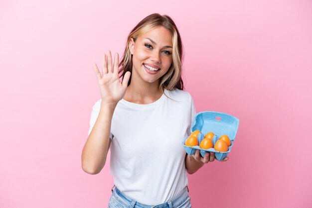Young Russian woman holding eggs isolated on pink background saluting with hand with happy expression