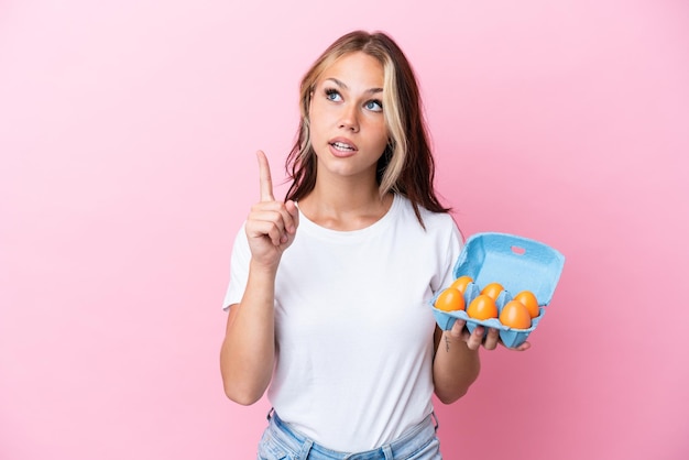 Young Russian woman holding eggs isolated on pink background thinking an idea pointing the finger up