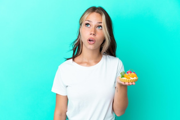 Young Russian woman holding a fruit sweet isolated on blue background looking up and with surprised expression