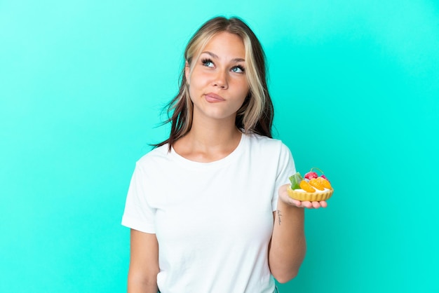 Young Russian woman holding a fruit sweet isolated on blue background and looking up