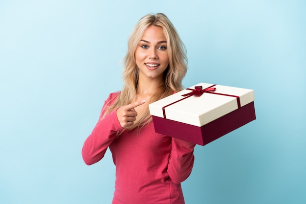 Young Russian woman holding a gift isolated on blue background and pointing it