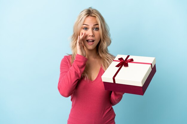 Young Russian woman holding a gift isolated on blue wall shouting with mouth wide open