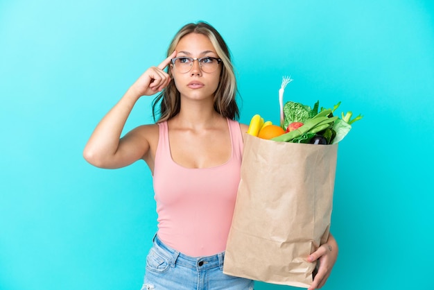 Young Russian woman holding a grocery shopping bag isolated on blue background having doubts and thinking