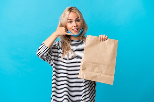 Photo young russian woman holding a grocery shopping bag isolated on blue background making phone gesture. call me back sign