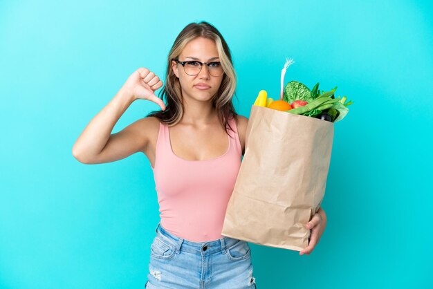 Young Russian woman holding a grocery shopping bag isolated on blue background showing thumb down with negative expression