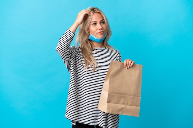 Young Russian woman holding a grocery shopping bag isolated on blue having doubts while scratching head