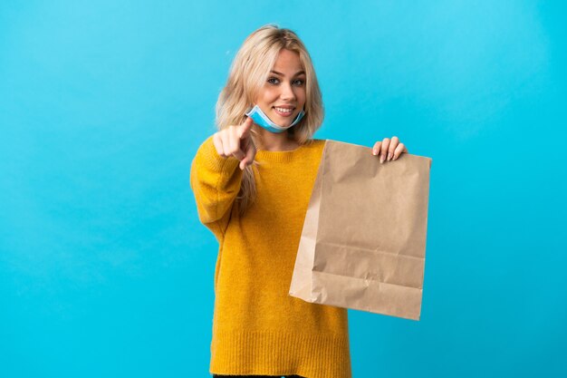 Young Russian woman holding a grocery shopping bag isolated on blue wall pointing front with happy expression