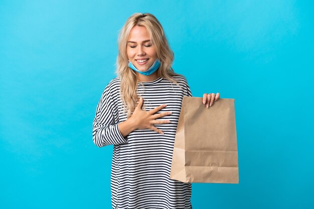 Young Russian woman holding a grocery shopping bag isolated on blue wall smiling a lot