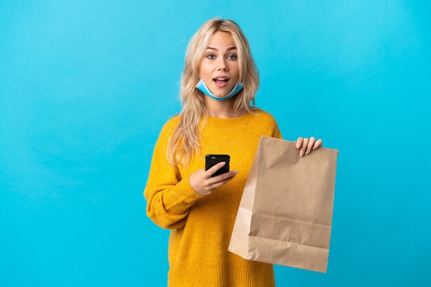 Young Russian woman holding a grocery shopping bag isolated on blue wall surprised and sending a message