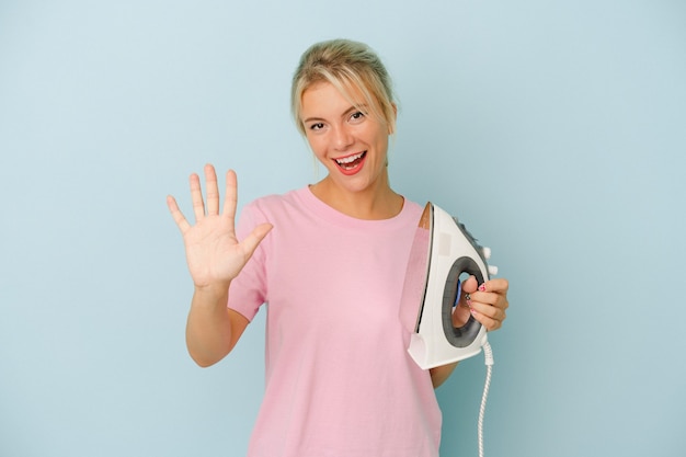 Young Russian woman holding iron isolated on blue background smiling cheerful showing number five with fingers.